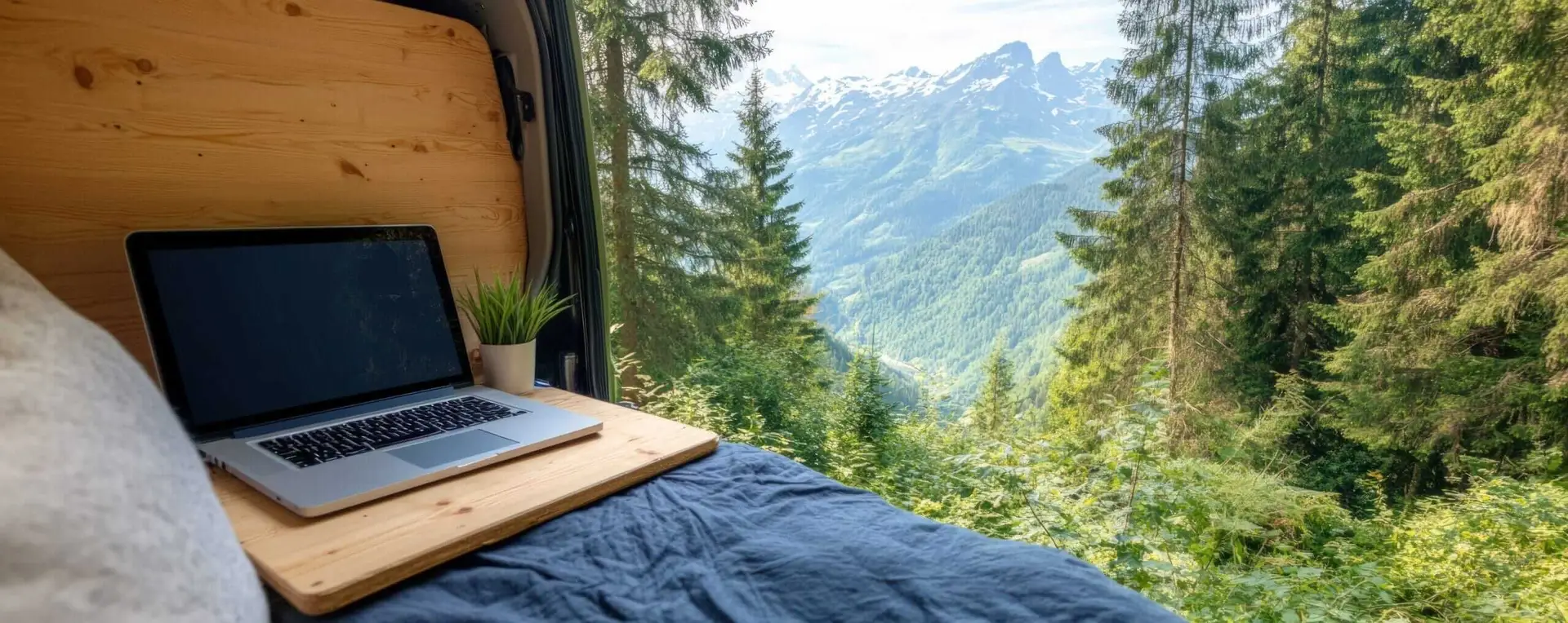 Bureau nomade avec ordinateur portable dans un van aménagé avec vue sur les montagnes et forêt de sapins.
